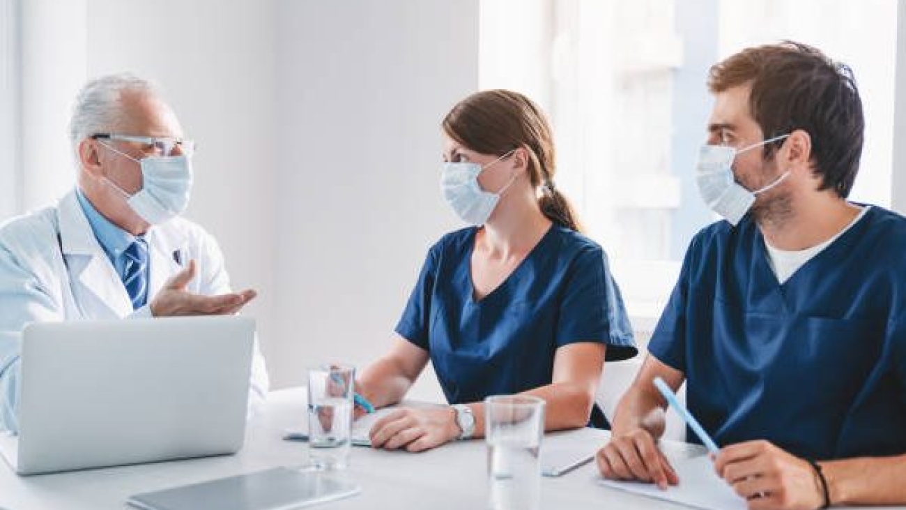 Senior medicine professional having meeting with his team in protective medical mask in boardroom during coronavirus